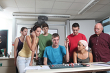 The picture shows a group of six young people and one adult looking at an experimental setup at a table. The young man in the middle is explaining something while the others listen with interest or interact with equipment.