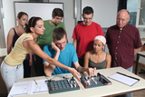 In the picture, several young people and a teacher are working together on experimental materials at a table. They seem to be concentrating on a technical task while discussing things with each other and exchanging instructions.