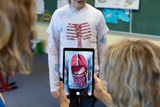 In the picture, a child is wearing a special printed T-shirt depicting the internal organs. A person is holding a tablet that displays an expanded representation of the organs, making learning about the human body interactive.