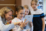 The picture shows a teacher and several students looking at a tablet together and interacting with each other. The atmosphere seems friendly and curious, which indicates a learning activity.