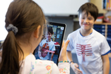 The image shows a girl using a tablet to take a picture of a boy wearing a special T-shirt that shows anatomical details. The boy is standing in front of her smiling and school supplies and the classroom are visible in the background.