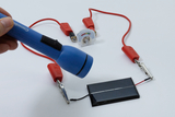 The image shows an experimental setup where a blue flashlight is pointed at a solar panel connected to red cables. In the middle is a meter for displaying electrical values, visualizing the connection and the functioning of the system.
