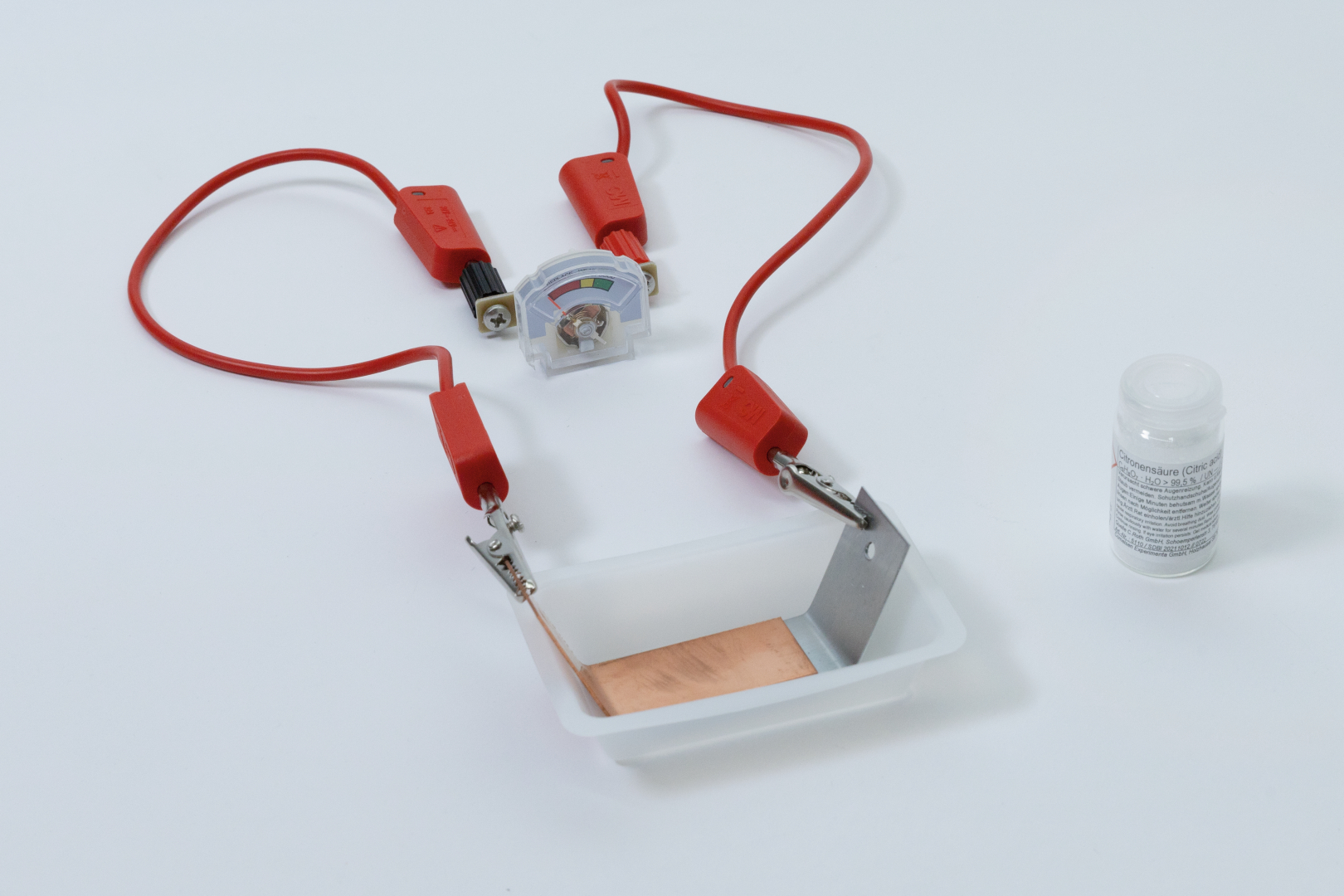 The picture shows an experiment set with two red cables, a multimeter for measuring voltage and a small plastic bowl equipped with a copper and a metal plate. Next to the set is a small glass of citric acid that can be used for chemical experiments.