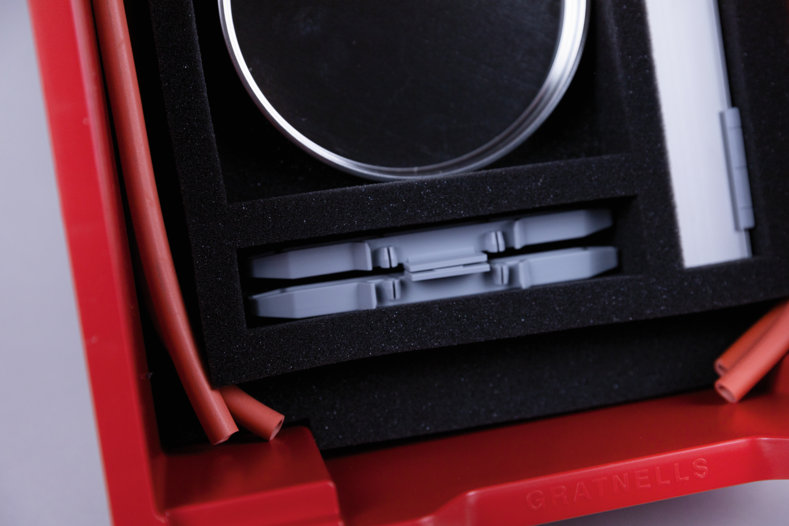 The picture shows a red storage box lined with black foam. Inside the box are various components including a glass container, grey parts and red tubing that can be used for experimental purposes.