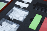 The image shows a transparent box with many small white building blocks neatly arranged on a foam base. Also visible are several colored plates and rubber tires, part of an educational experiment kit.