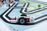 The image shows a toy vehicle made of modular building blocks, placed on a play mat with a track. The math is colorfully designed and features various symbols and QR codes that can be used for experiential learning.