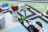 The image shows a scene from an educational toy consisting of modular building blocks representing a robotic arm and a vehicle. The robotic arm grasps a green block while the scene unfolds on a colorful, playful game board.