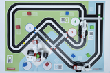 The image shows a play mat with a road layout, on which various stations and colored blocks are placed. Two robot vehicles made of building blocks are positioned on the mat, ready to explore the route.