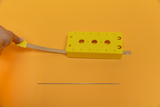 The image shows a yellow experimental device with a flexible metal tab and a straight metal rod lying on an orange background. The device appears to be designed for educational purposes and could be used for physics experiments.