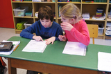 The picture shows two children sitting at a table, looking attentively at worksheets. The setting is a classroom atmosphere, with various materials and a bookshelf in the background.