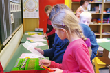 The picture shows a group of children in a classroom using an experiment station. Two children are looking at instructions together, while in the background other students and a teacher can be seen working at a table.
