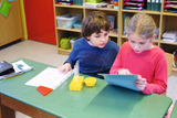 The picture shows two children sitting at a table in the classroom, working with experiment materials, including test tubes and a worksheet, while one of them looks at a tablet.