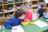 The picture shows two children working at a table with experimental materials. They are concentrating on a yellow set of test tubes and seem interested in a scientific experiment.