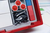 The picture shows an organizer with various experimental materials, including three round containers with red heads and a bag with black components. In addition to these, there are also colored plastic bricks in blue and red.
