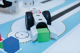 The image shows a technical model consisting of a vehicle with large rubber tires and a green cube component. It stands on a colored background with various geometric shapes and lines that apparently serve a learning or experimental scenario.