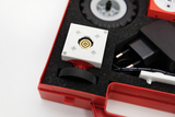 The picture shows an experimental kit arranged on foam padding in a red case. The focus is on a white module with a central mechanism, surrounded by other components such as tires and a power supply.
