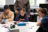The picture shows four students curiously looking at an experiment kit with a small robot. They are sitting at a table surrounded by school materials while a teacher stands in the background.