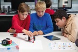 In the picture, three children are working concentratedly at a table while tinkering with a small, homemade robot. There are also some books and tablets on the table to support the experimentation.