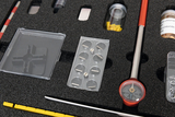 The picture shows a set of experimental supplies that includes various transparent containers, a measuring device with a red housing, and some small tools and containers. The arrangement of the parts is kept in a foam-padded box that offers protection and clarity.
