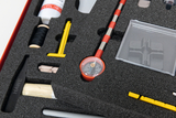 The picture shows a selection of experimental materials stored in structured foam. Various tools and materials can be seen, including a measuring device, a container and various writing and craft utensils.