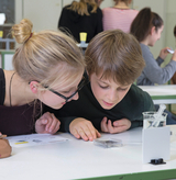 The picture shows two children looking curiously over a tabletop while concentrating on an experiment. In the background you can see other students who are also working with experimental materials.