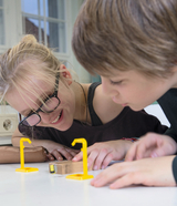 The picture shows two children looking intently at a small wooden block placed between two yellow experiment stands. They seem excited and curious as they examine the experimental materials.