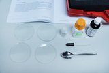 Four glass dishes are arranged neatly on a table, surrounded by various chemicals and laboratory equipment, including a spoon, a pipette, and a container of sugar. In the background, an instruction manual is visible, providing instructions for experiments.