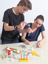 Two students are conducting an experiment in which they transfer a sample into a test tube using a pipette. Various fruits and vegetables, as well as chemical utensils such as test tubes and measuring beakers, are arranged on the table.