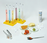 Six test tubes filled with colorful liquids sit in a yellow holder. Next to them are various pieces of fruit on Petri dishes, a tablespoon of an orange substance, and test strips for experiments.