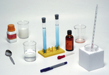 Various laboratory equipment and chemicals for experiments are arranged on a white background. You can see test tubes with blue solution, measuring beakers, pipettes, spoons, and bottles with clearly labeled substances.