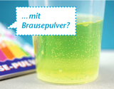 In the foreground, a transparent beaker containing a bubbling, greenish liquid is visible. In the background, a colorful package of effervescent powder lies, suggesting possible experimental instructions.