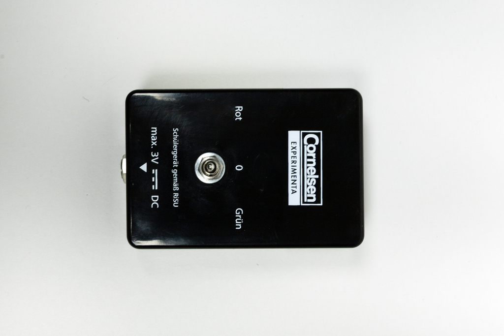 A black experiment device features a connector socket and colored labels in red and green. It is designed for use in schools and supports a maximum voltage of 3 volts DC.