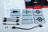 On the table are several electronic components in gray cases displaying various circuit symbols. There are also some cables with connectors and two light bulbs, as well as an open manual with circuit diagrams for explanation.