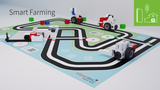 Eine Spielmatte mit einem kreisförmigen Straßenmuster dient als Basis für eine Smart Farming-Lernumgebung. Auf der Matte befinden sich verschiedene bunte Bausteine und mobile Roboterfahrzeuge, die interaktive Experimentiermöglichkeiten bieten.