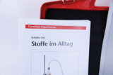 Ein Schüler-Set namens „Stoffe im Alltag“ von Cornelsen Experimenta liegt im Vordergrund. Dahinter ist ein Teil eines roten Transportkoffers mit schwarzem Schaumstoff zu sehen.