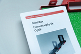 Ein Heft mit dem Titel „Mini-Box Elementarphysik. Optik“ von Cornelsen Experimenta liegt auf einer hellen Fläche. Im Hintergrund ist ein Teil einer roten Box mit grünem Schaumstoff zu sehen.