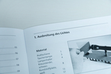 Ein aufgeschlagenes Lehrbuch zeigt eine Anleitung zur „Ausbreitung des Lichtes“. Unter der Materialliste ist ein Foto zu sehen, das den Versuchsaufbau mit einer Taschenlampe und einer Kunststoffplatte illustriert.