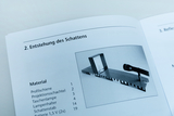 Ein offenes Heft zeigt eine Anleitung zur „Entstehung des Schattens“. Eine Materialliste führt unter anderem eine Profilschiene, eine Projektionsschachtel und eine Taschenlampe auf.