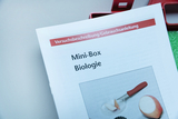 Eine Bedienungsanleitung für eine „Mini-Box Biologie“ liegt auf einer hellen Fläche. Das Cover zeigt neben dem Titel ein Foto mit einem Präpariermesser, einem Objektglas und einem Apfelstück.