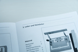Eine geöffnete Broschüre zeigt das Kapitel „Leiter und Nichtleiter“ für physikalische Experimente. Neben einer Materialliste ist eine Abbildung eines Versuchsaufbaus mit Stecksockeln und Schaltern zu sehen.