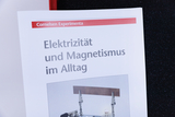 Gezeigt wird eine Broschüre mit dem Titel „Elektrizität und Magnetismus im Alltag“ aus der Reihe Cornelsen Experimenta. Unter dem Text ist ein Teil eines physikalischen Versuchsaufbaus zu sehen.