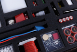 Verschiedene physikalische Experimentierkomponenten liegen sortiert in einem schwarzen Schaumstoffinlay. Zu sehen sind unter anderem rote Kabel, Batterien, elektrische Bauteile und kleine Zubehörteile in Kunststoffboxen.