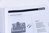 Ein aufgeschlagenes Lehrbuch zeigt unter der Überschrift „Elektrizität“ den Aufbau eines elektrischen Stromkreises. Neben einer schematischen Abbildung ist eine Materialliste mit benötigten Komponenten wie Glühlampenfassung und Batterien aufgeführt.