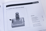 Eine Anleitung zur Funktion einer elektrischen Klingel zeigt einen Versuchsaufbau auf einem Stecksockel. Rechts daneben ist die benötigte Materialliste für das Experiment aufgeführt.