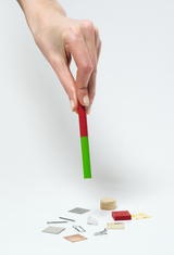 Eine Hand hält einen rot-grünen Stab über verschiedene kleine Materialproben. Auf der weißen Fläche liegen Metallplättchen, Holz, Kunststoff und andere Testobjekte.
