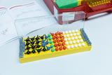 Ein gelber Molekülbaukasten mit farbigen Kunststoffkugeln und Verbindungsstücken liegt geöffnet auf einer weißen Fläche. Im Hintergrund sind ein roter Koffer sowie ein aufgeschlagenes Heft mit chemischen Strukturformeln zu sehen.