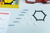 Ein Chemie-Molekülbaukasten mit grauen Verbindungsstücken, weißen Atomsphären und einem schwarzen Sechserring liegt auf einer weißen Fläche. Daneben ist ein Teil eines Anleitungsblatts mit chemischen Formeln zu sehen.