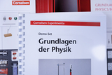 Ein spiralgebundenes Heft mit dem Titel „Grundlagen der Physik“ aus dem Cornelsen Experimenta Demo-Set steht im Vordergrund. Im Hintergrund sind Teile von Physik-Experimentierkästen erkennbar.