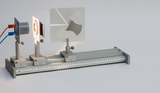 Ein optisches Experimentierset auf einer metallischen Schiene zeigt eine Lichtquelle und einen Bildschirm. Die Anordnung dient der Demonstration physikalischer Lichtphänomene im Unterricht.