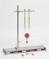 Ein Physik-Experimentierset zeigt zwei Federwaagen, die an einem Stativ hängen und über eine rote Rolle verbunden sind. Drei kleine Zusatzgewichte liegen davor auf einer markierten Basisplatte.